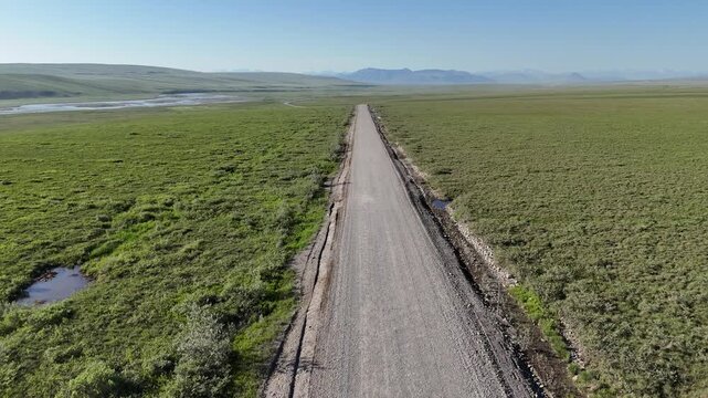 Aerial perspective flying over the endless dalton highway, a remote gravel road stretching through the vast green tundra and permafrost landscape of northern alaska towards distant mountains