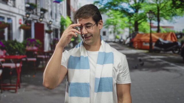 Man adjusts glasses while wearing a blue and white striped scarf and white tshirt in a street cafe with tables and trees visible; content.