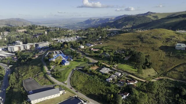 Drone flies south high over building with blue roof on hill next to highway in the late afternoon before sunset in mountain town of Mbabane, Eswatini