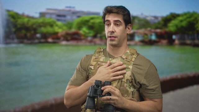 Man in camouflage vest and tactical uniform holds binoculars then places hand on chest on a street walkway; determination duty.
