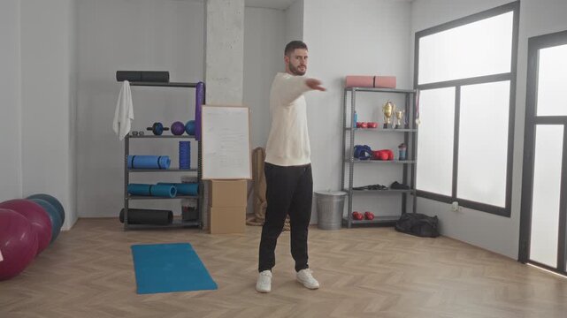 Man with arms outstretched performing a standing balance yoga pose on mat in a fitness studio, visible exercise gear and shelves nearby; calm balance.