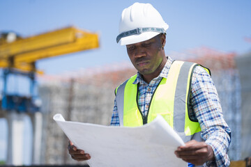 african american civil engineer reviewing blueprints at a construction site, professional construction worker in a hard hat and safety vest holding floor plans © manit