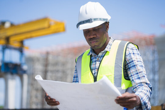 african american civil engineer reviewing blueprints at a construction site, professional construction worker in a hard hat and safety vest holding floor plans