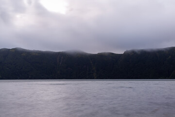 Fototapeta premium Sete Cidades volcanic lake in San Miguel Island, Azores, Portugal. Calm water reflects cloudy sky. Lush green hills surround the crater lake.