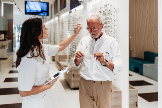 An elderly man receives professional help from a female optician while choosing and fitting new glasses in a bright, modern optical shop.