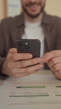 Man holding smartphone, fingers tapping screen above printed invoice and charts on table in building; concentration productivity.