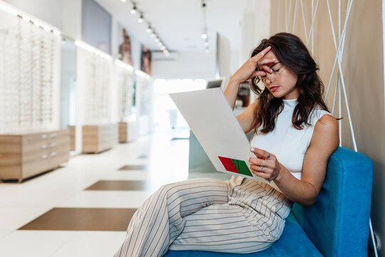 A focused young woman sits on a blue sofa in a modern optical store, holding a vision test chart and adjusting her new eyeglasses to check her sight.