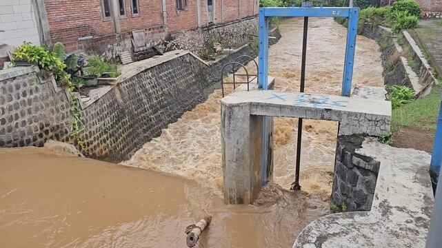 Fast-flowing, muddy floodwater rushes past a concrete and blue metal floodgate in a stone-lined urban canal following heavy rainfall, threatening the nearby residential embankments.