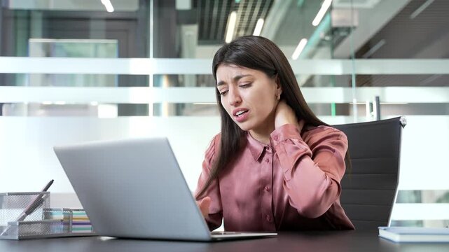 Businesswoman suffering from neck pain while working at desk in modern office, massaging sore muscles. Tired female employee easing discomfort, loosening stiff neck, coping with fatigue in workplace.