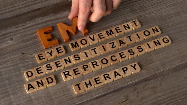 A hand arranging the abbreviation EMDR with wooden letters.  Its explanation: Eye Movement Desensitization and Reprocessing, formed with wooden letter tiles, is visible. Rustic wooden background.  