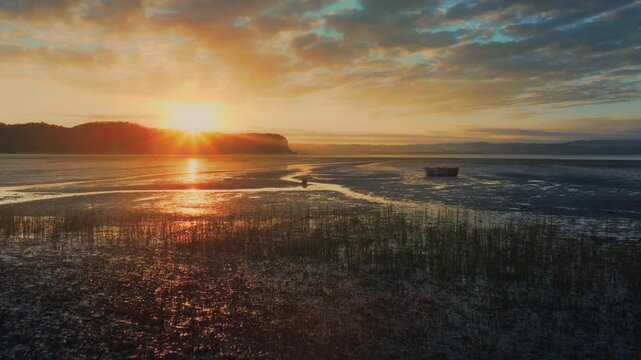 A lone pontoon rests on a mudflat during a vibrant sunset at Kawhia Harbour, Kawhia, Waikato, New Zealand. The sun's rays pierce through the clouds, casting a warm glow over the tranquil landscape.