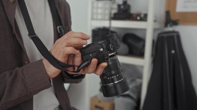 Man adjusting camera with hands and strap in studio, photographer checking lens, aperture and controls while standing near shelving and gear; quiet concentration.