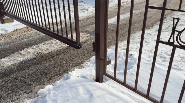 Snow covered driveway iron gate opening into residential parking lot gravel tracks and compacted snow mark vehicle path rust brown.