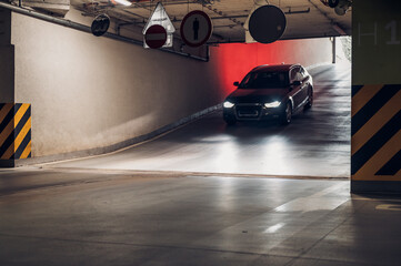 Naklejka premium Car exits underground parking garage in urban area during evening hour with signs visible and concrete surface illuminated by headlights