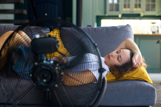 Woman sleeping on couch with fan during heatwave. Summer fatigue concept