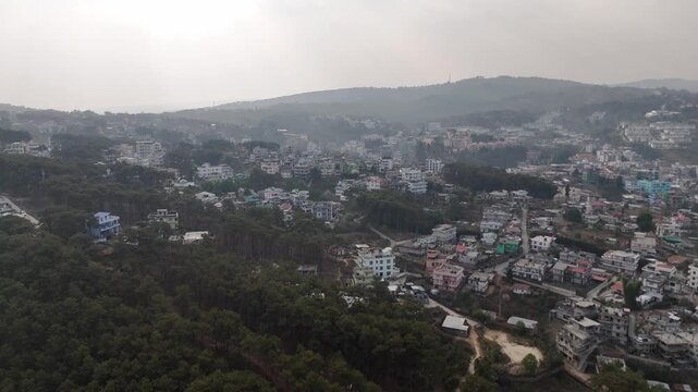 Drone footage captures a misty hillside town with colorful multi-story buildings scattered across green slopes. Dense pine forest covers the foreground while residential structures in white pink and b