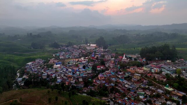 Drone shot capturing Valparai at sunset, featuring dense housing clusters, winding roads, green hills, and soft orange light breaking through clouds over layered mountains.