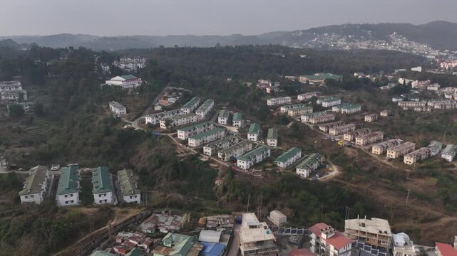 Drone footage captures hillside residential colony featuring white buildings with distinctive green rooftops spread across terraced slopes surrounded by dense forest and misty mountains in the backgro