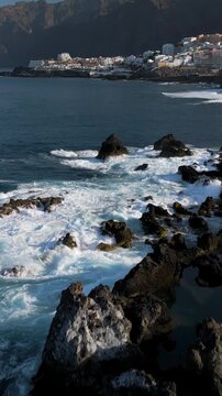Aerial view of foamy sea waves breaking on the rocky coast of Los Gigantes. Panorama of the city of Puerto de Santiago, Playa de la Arena and Los Gigantes, Tenerife, Canary Islands, Spain