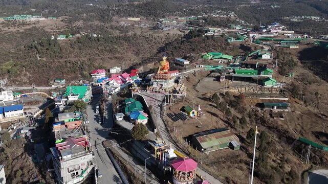 Drone footage reveals a Himalayan mountain town with colorful rooftops surrounding a golden Buddha statue. Winding roads connect buildings with green and red metal roofs across the rugged hillside ter