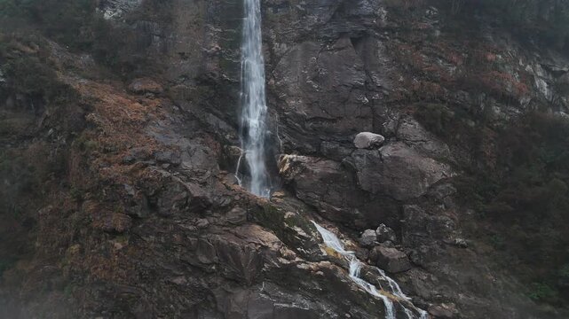 Majestic waterfall plunges down rugged dark rocky cliff face with misty spray. Water flows over moss-covered brown and grey rocks surrounded by sparse vegetation. Moody overcast atmosphere creates dra