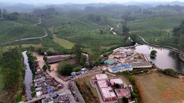 Cinematic aerial view of Valparai showcasing a serene hill station lake surrounded by tea plantations, winding streams, and scattered buildings, with lush greenery stretching across rolling hills.