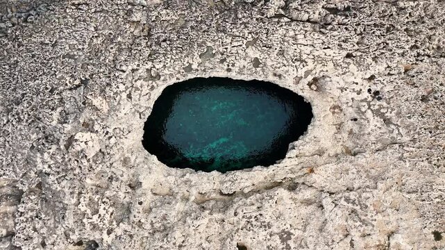 Blue water sits inside a rounded hole in pale limestone rock at Coral Lagoon, Malta, showing a natural opening in a close overhead view with textured stone around it.