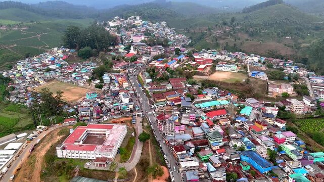 Wide cinematic aerial of Valparai highlighting vibrant townscape, layered hills, plantation patterns, and a moody sunset sky creating depth and atmosphere.