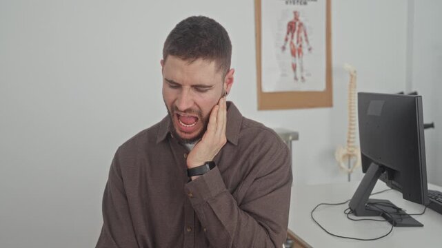 Man touching jaw with hand and wincing, hand on cheek and eyes closed, seated at desk by monitor and anatomy poster in building clinic; pain concern.
