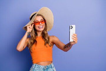 Young chic blonde woman taking a selfie with hat and sunglasses smiling against blue background