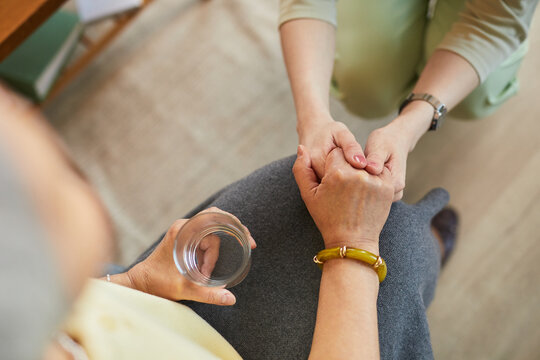 Senior woman receiving supportive handhold from female caregiver in nursing home, holding drinking glass
