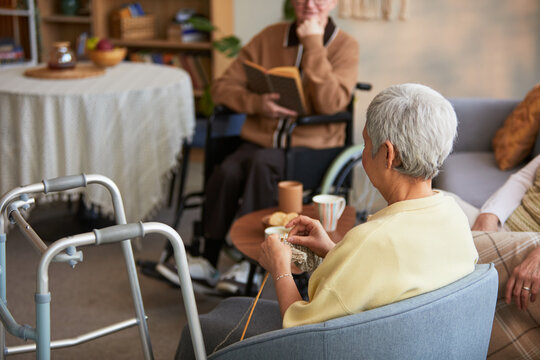 Senior women socializing in nursing home, reading book and knitting near mobility aids, representing elder care and assisted living