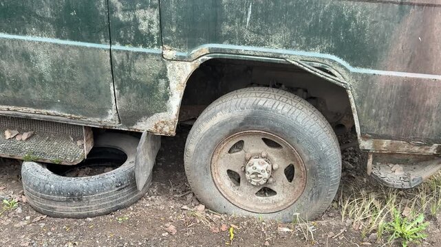Abandoned dark green minibus punctured tire, rusted undercarriage and chipped paint, spare tires stacked beneath body, moss.