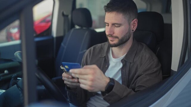 Man in car holding creditcard and smartphone, hands tapping phone while seated on street; focused mobile payment.