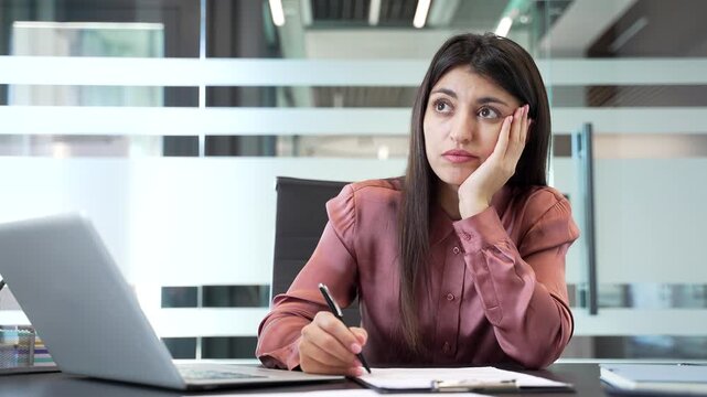 Bored businesswoman sitting at desk in office with pen and documents. Female employee feeling uninterested and tired, doing routine paperwork, representing monotony and lack of motivation in workplace