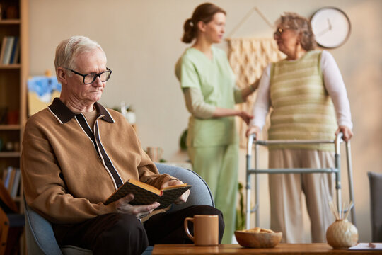 Senior Caucasian man reading book in nursing home while caregiver assisting senior woman with walker, showing elder care support
