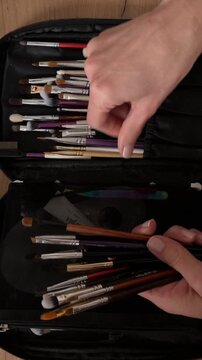 Artist organizing paint brushes in black case top view. Top down close up of female hands sorting assorted paintbrushes into elastic loops of a black portfolio case on a wooden desk, organizing art su