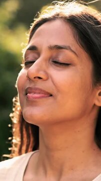 Closeup of a beautiful young Indian woman with her eyes closed, gently smiling, turning her face upwards to feel the warm, soft sunlight on her skin in serene outdoor video footage.