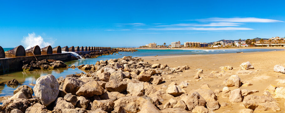 Playa de Cubelles, Catalu&ntilde;a, Espa&ntilde;a, costa mediterr&aacute;nea. Playa rocosa y arenosa con rompeolas, olas rompiendo, mar azul en calma y edificios costeros bajo un cielo brillante.