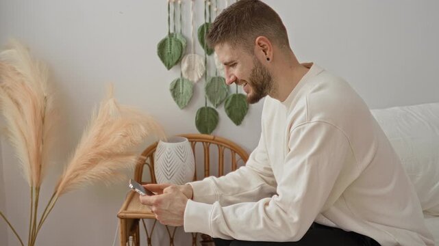 Man holding smartphone in both hands and smiling while sitting on bed beside rattan chair with pampas and vase in studio; quiet focused contentment.