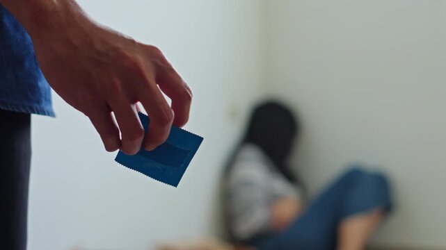 Close up of hand holding blue condom packet while woman sits distressed in background