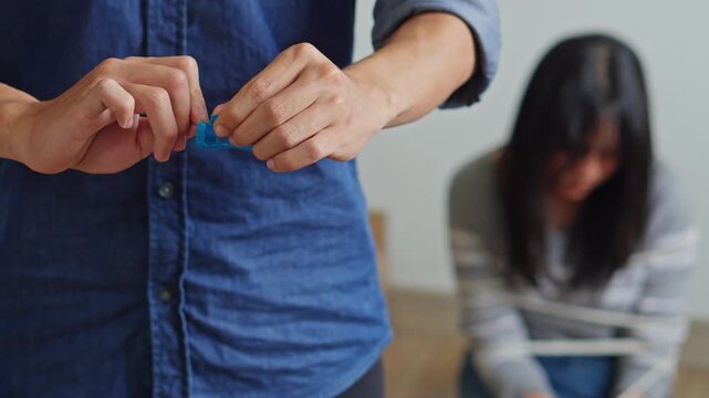 Close-up of trafficker hands opening condom wrapper with blurred tied-up woman in background