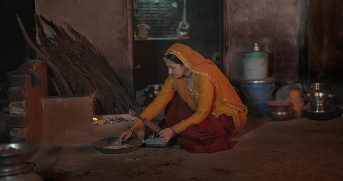 Indian mature adult chef woman mom wear saree sitting do work cook local food at dark room rural area. Female lady use metal chimta make fresh roti chapati on open fire flam clay stove home place