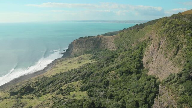 A scenic view of the rugged coastline and lush green hills of the Raglan, Waikato, New Zealand, with the turquoise ocean waves crashing against the shore.