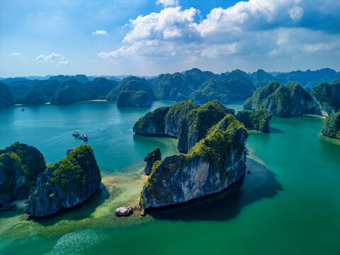 Aerial view of the limestone karsts and emerald waters of the Cat Ba Archipelago with small boats under a bright blue sky in Ha Tu, Hai Phong, Vietnam.