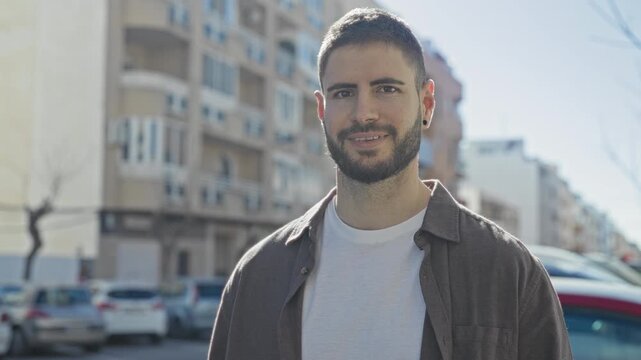 Man with earring and white t shirt smiles broadly while standing near parked cars and apartment buildings on a sunny street; warmth casual confidence.
