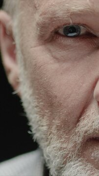 Vertical close view of half face of senior Caucasian man showing eye and skin details. Male showing skin texture and wrinkles. Looking forward with calm expression. Standing in dark studio background.