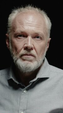 Senior Caucasian man looking at camera with serious expression. Male keeping steady gaze and calm face. Showing wrinkles and life experience. Standing in dark background in studio.