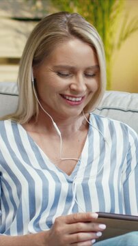 Caucasian woman wearing earphones and looking at smartphone with smile. Female listening to audio and enjoying content. Holding device in hands and relaxing. Sitting on sofa at home.