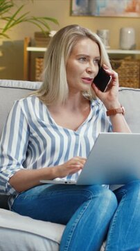 Caucasian woman holding smartphone and using laptop while sitting on sofa. Female talking during call and checking screen. Managing tasks and discussing details. Spending time at home.